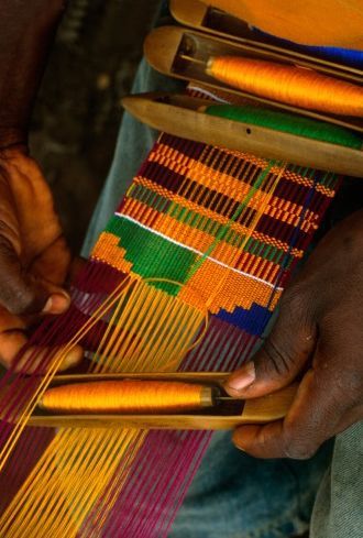 Hands weaving a strip of kente on a loom, shuttles of gold and green thread beside it